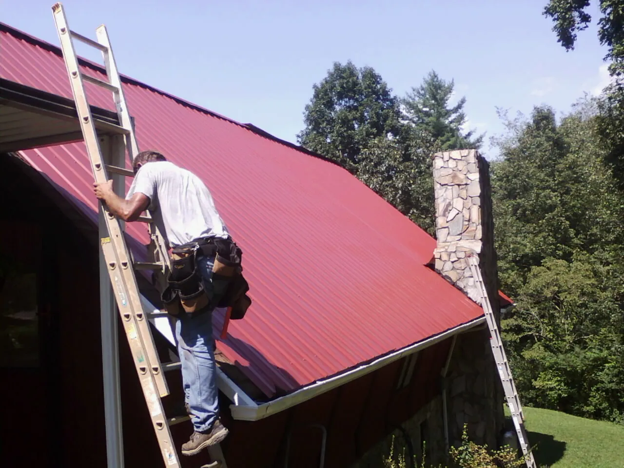 Roofer on a ladder beside a red metal roof and stone chimney — Soffit & Fascia Repair by Roofing Brothers in Eastlake Weir